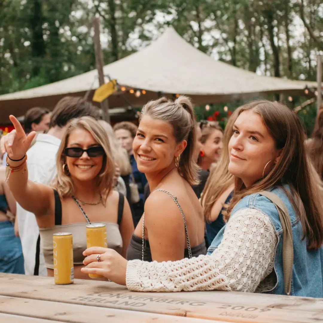 Een vrolijk moment vastgelegd terwijl drie vrouwen ontspannen aan een picknicktafel, genietend van drankjes en de levendige sfeer van het Daycare Festival in de Spoorzone van Tilburg.